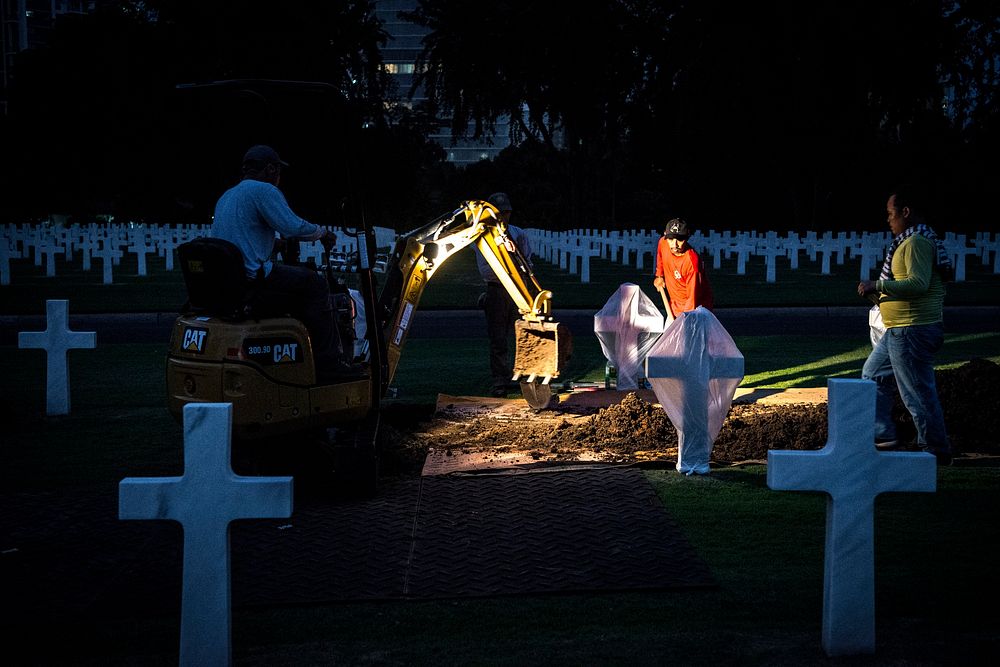 Local workers with the American Battle Monuments Commission work with members of the Defense POW/MIA Accounting Agency…