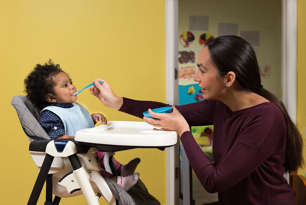 Child care provider sitting directly in front of an infant and using a spoon to place a small amount of baby food between…