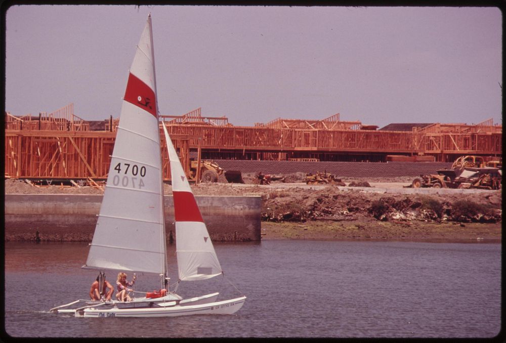 Sailboat glides past Sea Harbour, a major development under construction at Huntington Beach, May 1975. Photographer:…