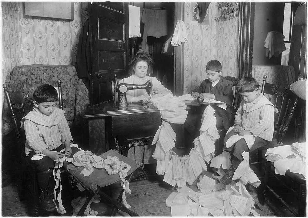 Making dresses for Campbell kids dolls in a dirty tenement. The older boy, about 12 years old, operates the machine when the…