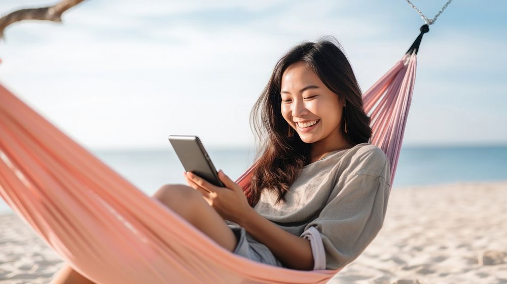 Happy traveler asian woman using mobile phone and relax in hammock on beach. AI generated Image by rawpixel.