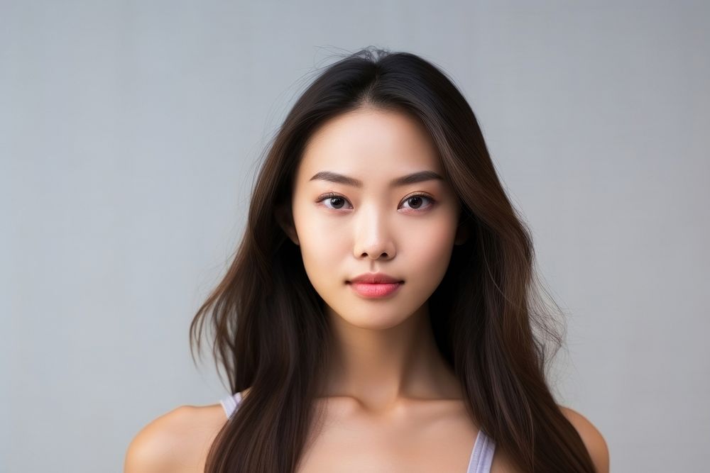 Portrait of a young woman with long dark hair, wearing a white top. She has a calm expression, standing against a neutral…