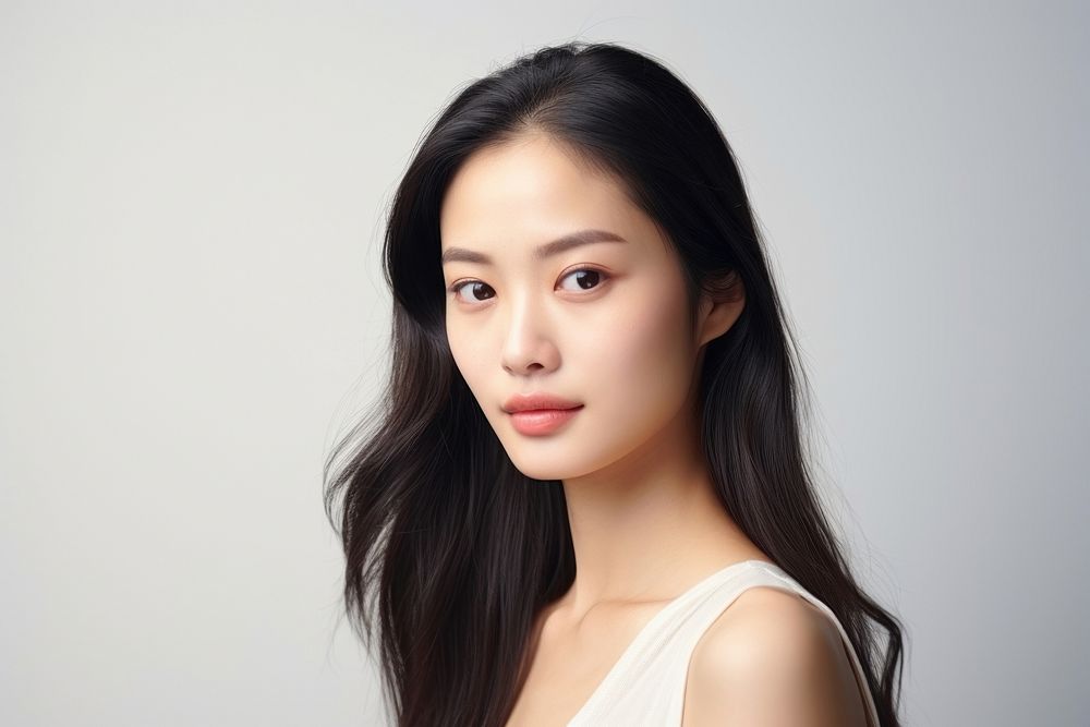 Portrait of a woman with long dark hair, wearing a white top, against a light background. She has a calm expression, looking…