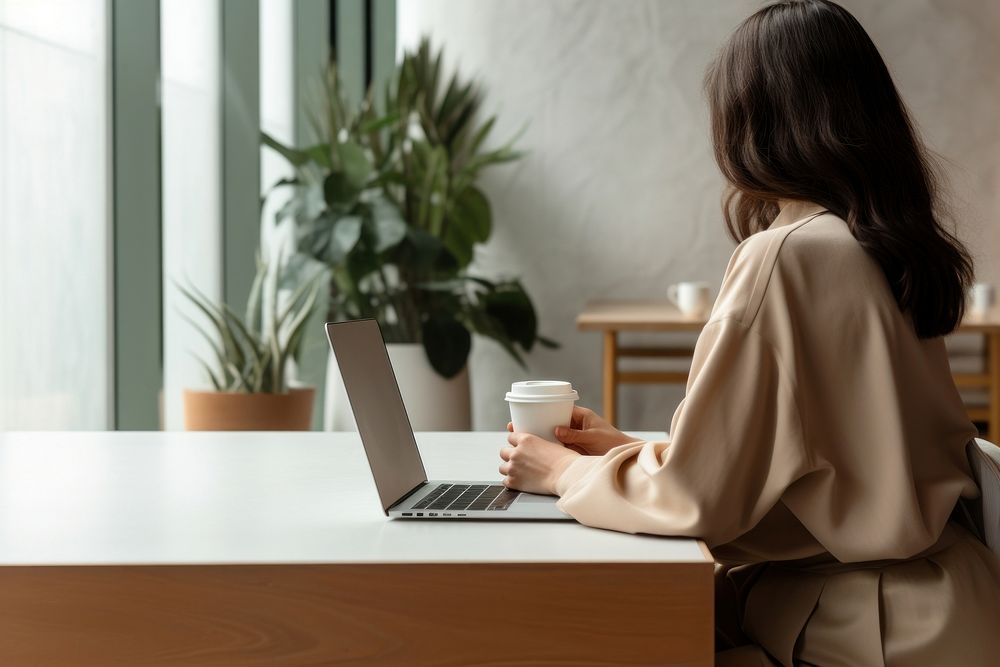 Woman sitting at a table with a laptop cup furniture computer. AI generated Image by rawpixel.