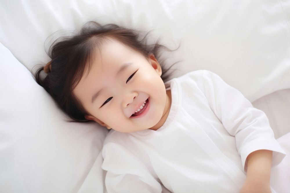 Smiling baby asian girl lying on a bed sleeping on white sheets smiling smile comfortable. AI generated Image by rawpixel.