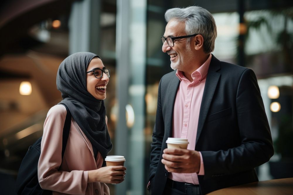 Woman in pink hijab and denim jacket talk to a cheerful CEO with long gray hair in brown sweater wearing eyeglasses scarf…