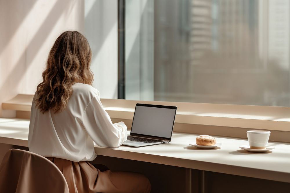 Woman sitting at a table with a laptop cup windowsill furniture. AI generated Image by rawpixel.
