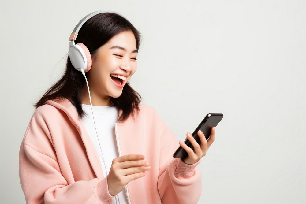 Asian teenage girl streaming online music on mobile phone headphones laughing headset. AI generated Image by rawpixel.