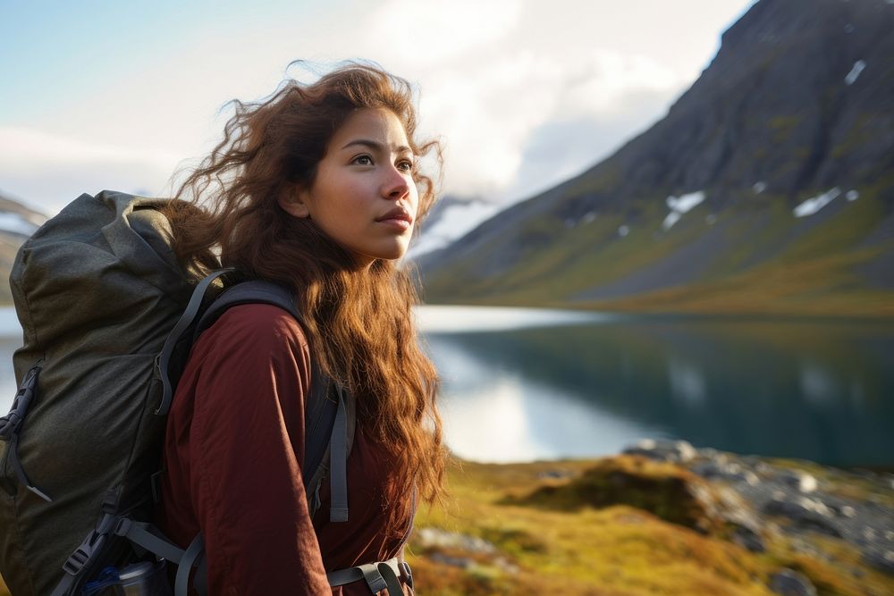 A Latina Brazilian teenage female backpacker looking at a reflective lake near a mountain in Greenland with the early…