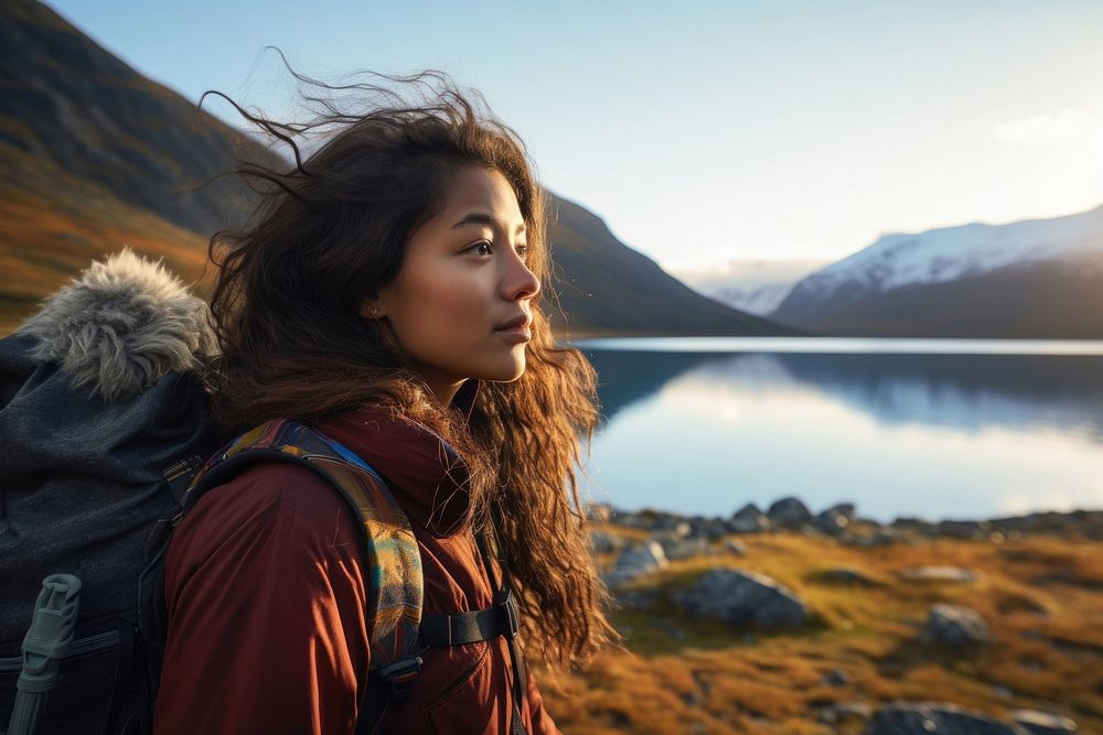 A Latina Brazilian teenage female backpacker looking at a reflective lake near a mountain in Greenland with the early…