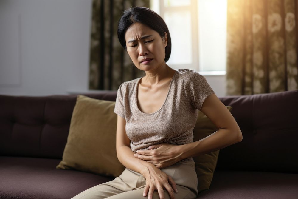 Asian middle age woman sitting on couch with stomach pain worried adult contemplation. AI generated Image by rawpixel.