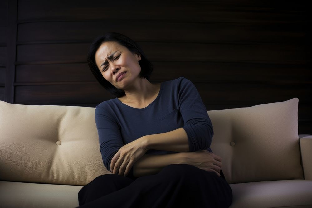 Asian middle age woman sitting on couch with stomach pain furniture portrait worried. AI generated Image by rawpixel.