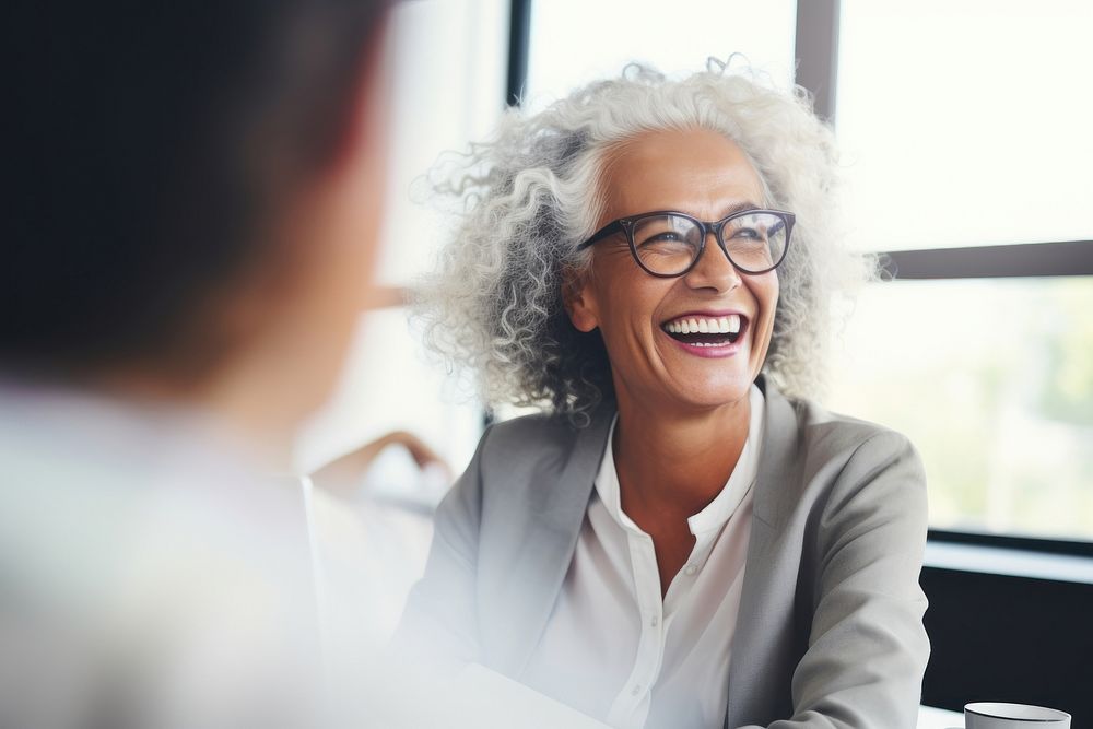 Mature businesswoman wearing eyeglasses laughing during meeting at office adult conversation togetherness. AI generated…