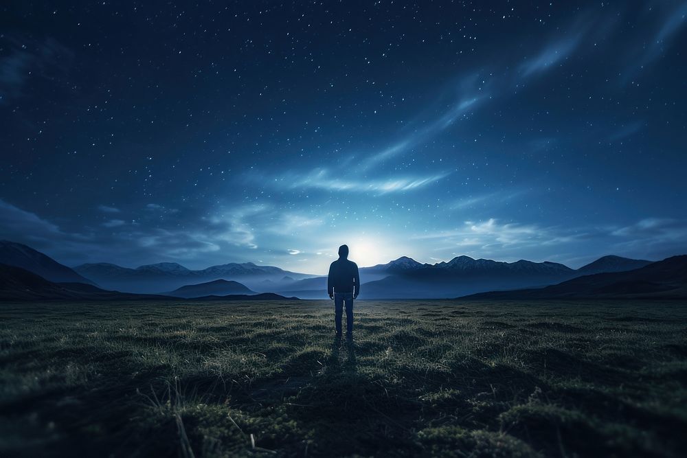 A man standing in a field by a starry night photography landscape mountain. AI generated Image by rawpixel.