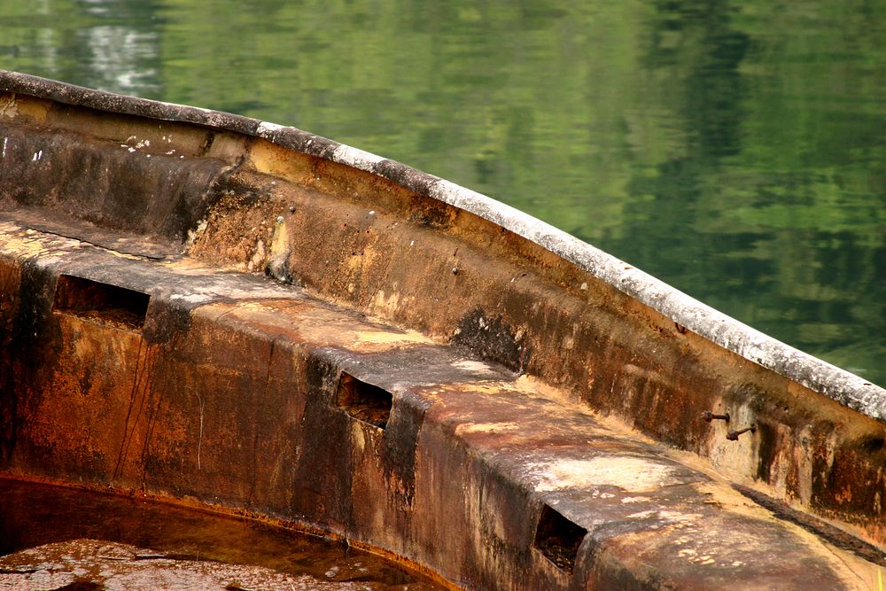 A large, curved, rusted metal edge leans over calm water reflecting shades of green.