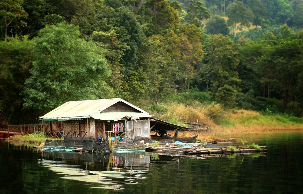 A shack on a river bed is surrounded by lush green jungled in the background. Some colorful shirts hang to dry outside.