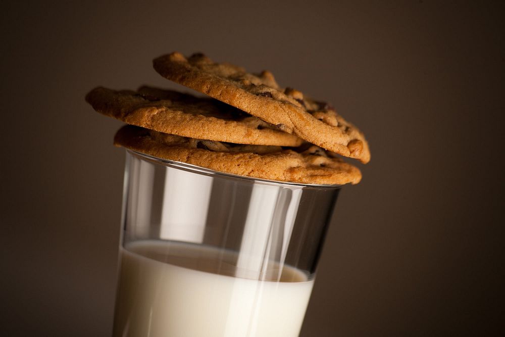 A close up of a glass of milk with a stack of three chocolate chip cookies precariously sitting on the rim of the glass.…