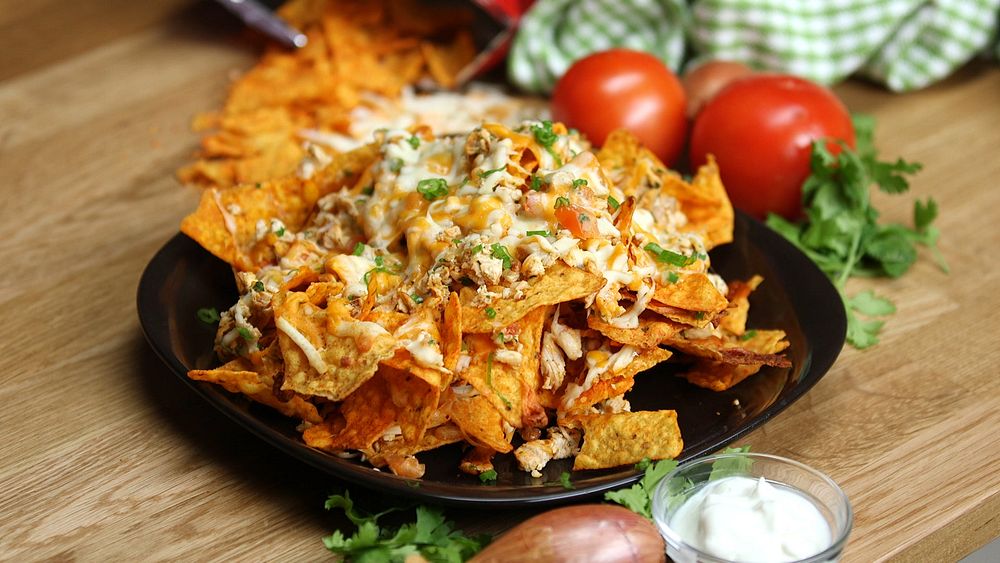 Plate full of nachos made with doritos topped with shredded chicken, green onions and tomato's on a table with a side of…
