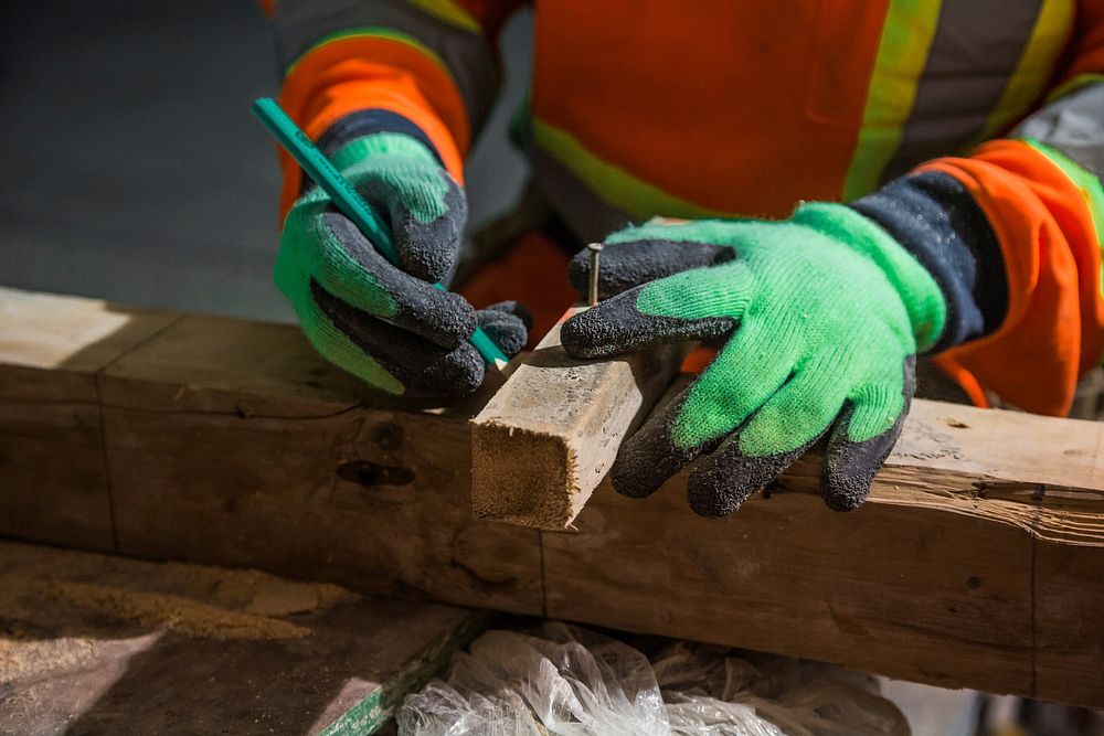 A construction worker in safety gloves measures wood before making a cut.