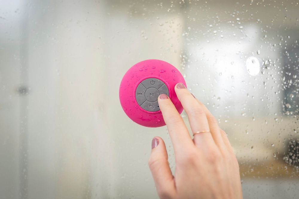 Woman finger pressing gray button on pink waterproof speaker in shower, public domain CC0 image.