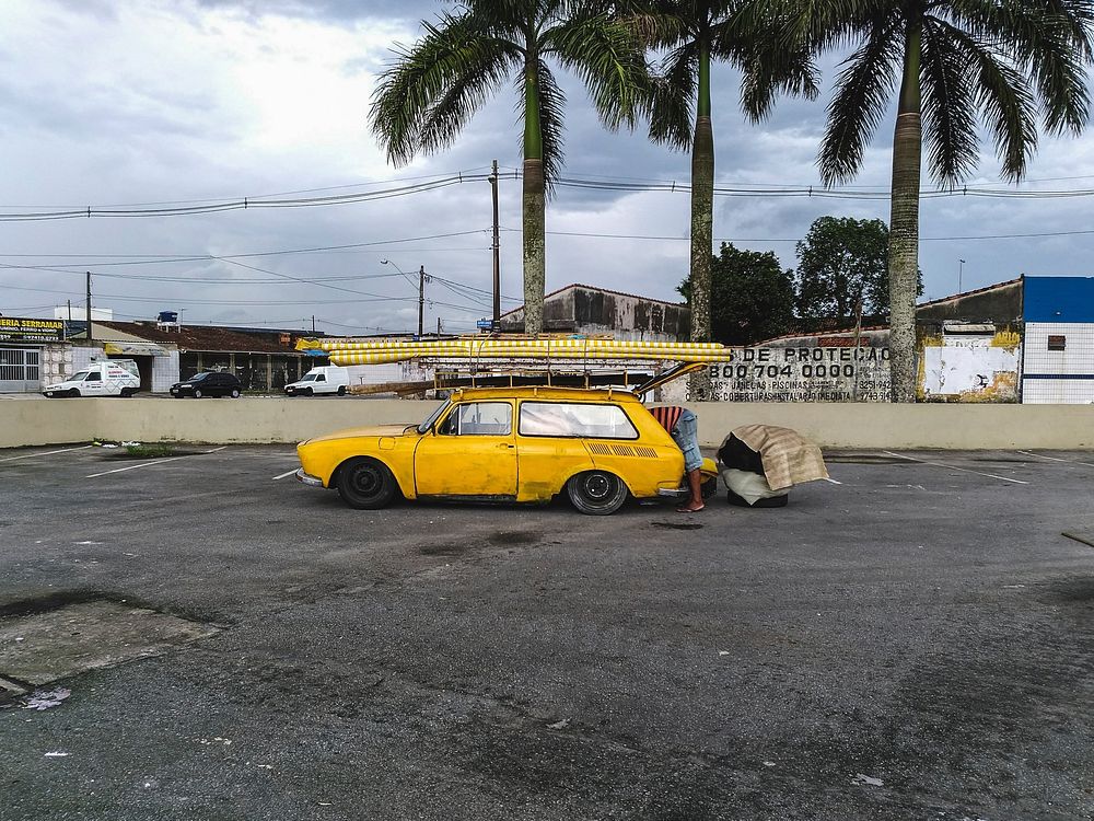 A yellow car with a loaded ski rack is parked in an empty lot in a tropical city. The driver is getting something out of its…