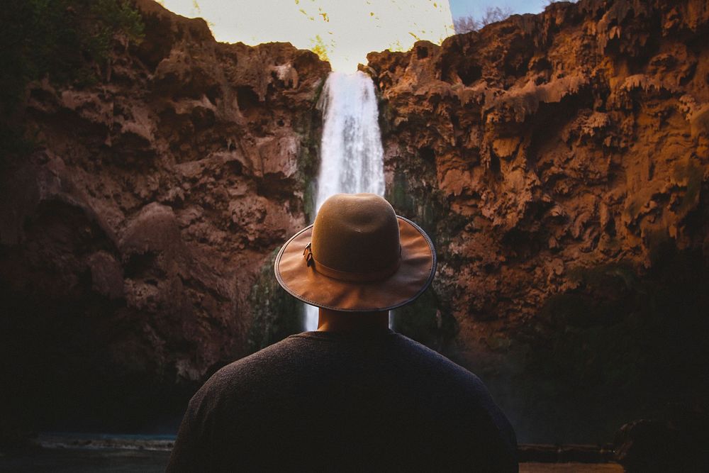 Free man with hat standing in front of a cliff waterfall photo, public domain CC0 image.