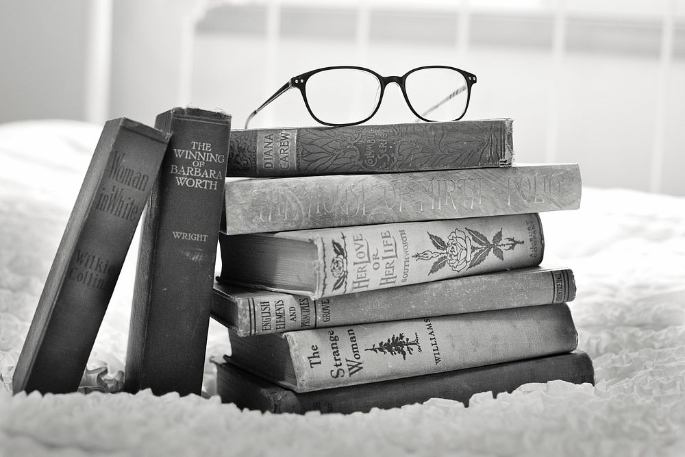 Free glasses on pile of books on bed in black and white photo, public domain CC0 image.