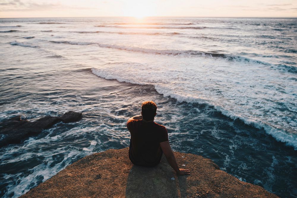 Man sitting on a cliff facing ocean view, free public domain CC0 photo.