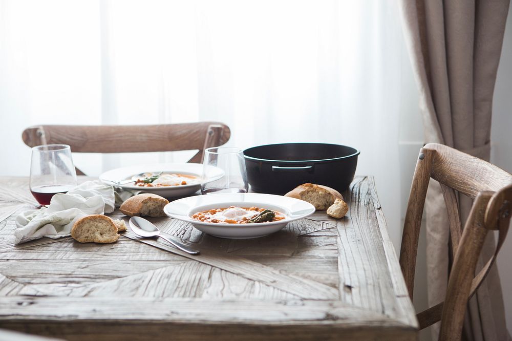 Two deep dishes with a hearty meal, pieces of bread and wine glasses on a wooden table. Original public domain image from…