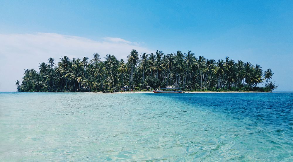 Distant view of a boat moored at an island covered with tropical trees. Original public domain image from Wikimedia Commons