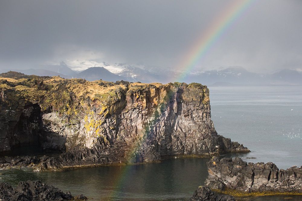 A rainbow crosses the center of the image, in front of a rocky cliff on water and a mountain range off in the distance.…