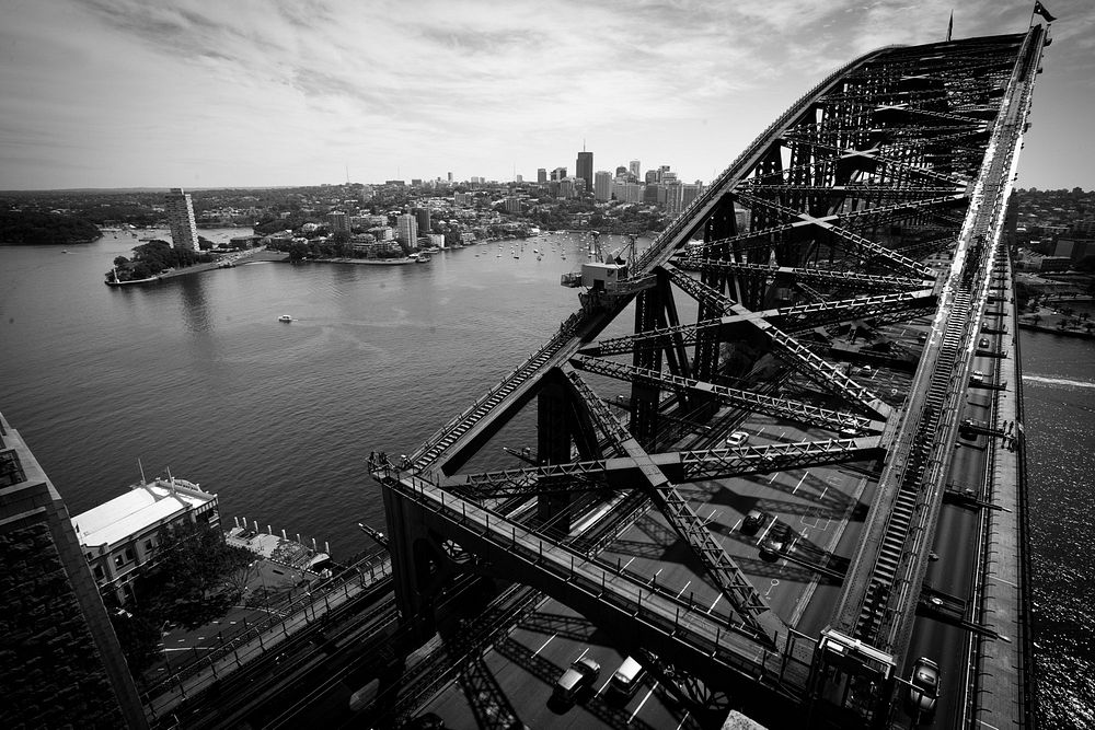 This black and white shot looking down at a bridge with cars driving across shows a busy harbor and city in the distance.