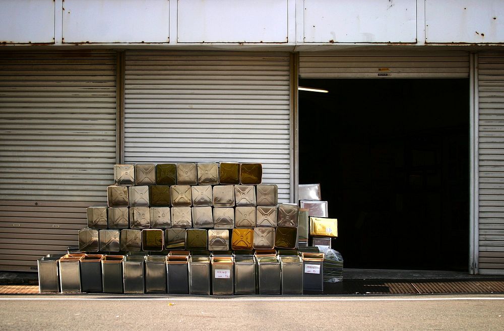 The sun glistens off a pile of metal tins, neatly stacked, outside the metal doors of a storage unit.