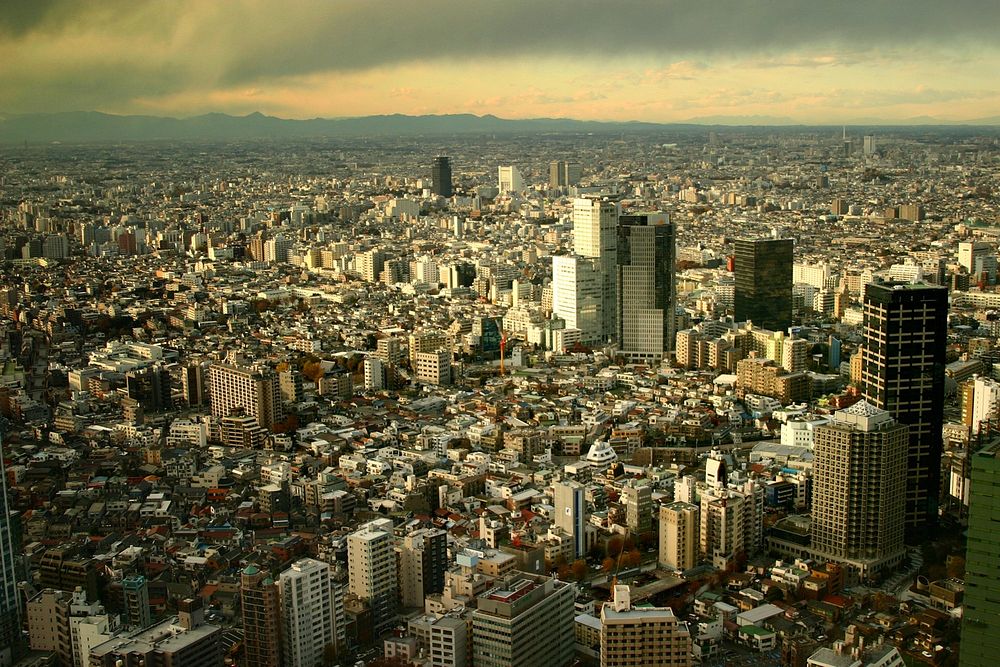 Shot from the air, this view of a densely packed Japanese city has mountains in the distance and dark clouds at the top of…