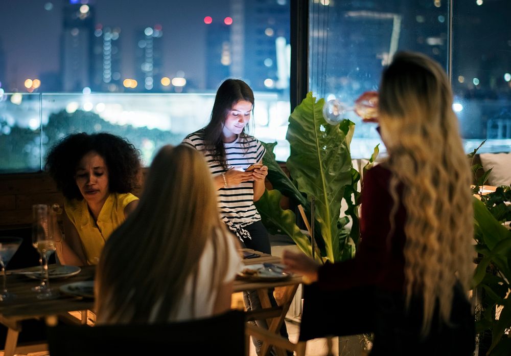 Young woman using a smartphone at a dinner night having no interaction with friends addiction concept