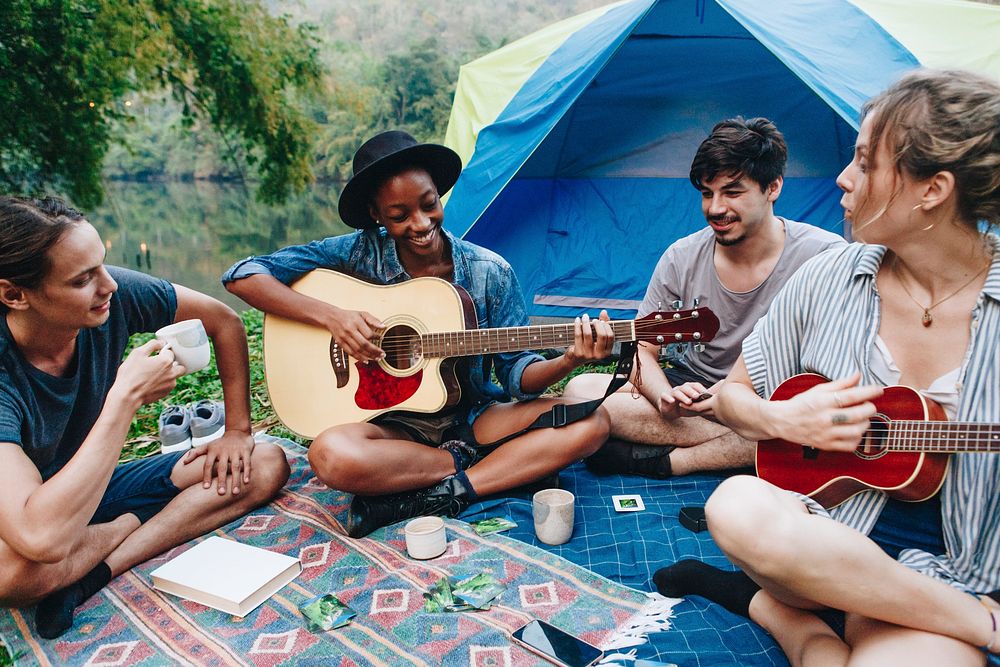 Group of young adult friends in camp site playing guitar and ukelele and singing together outdoors recreational leisure and…