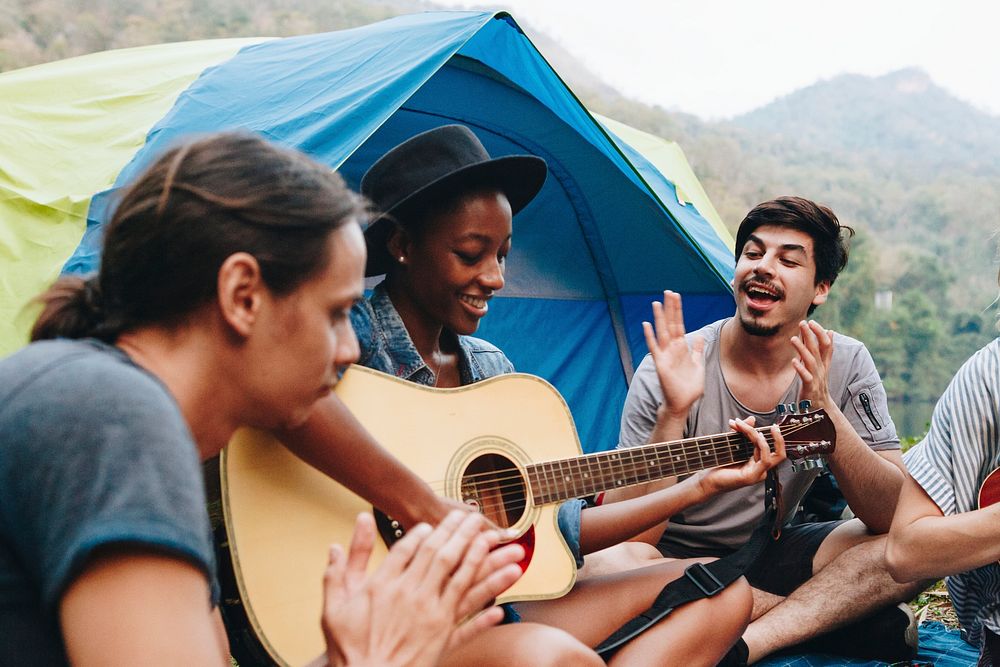 Group of young adult friends in camp site playing guitar and ukelele and singing together outdoors recreational leisure and…