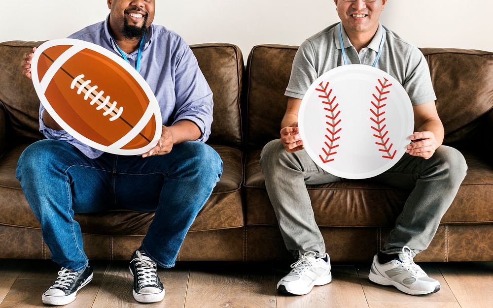 Men holding baseball and rugby icons sitting on couch