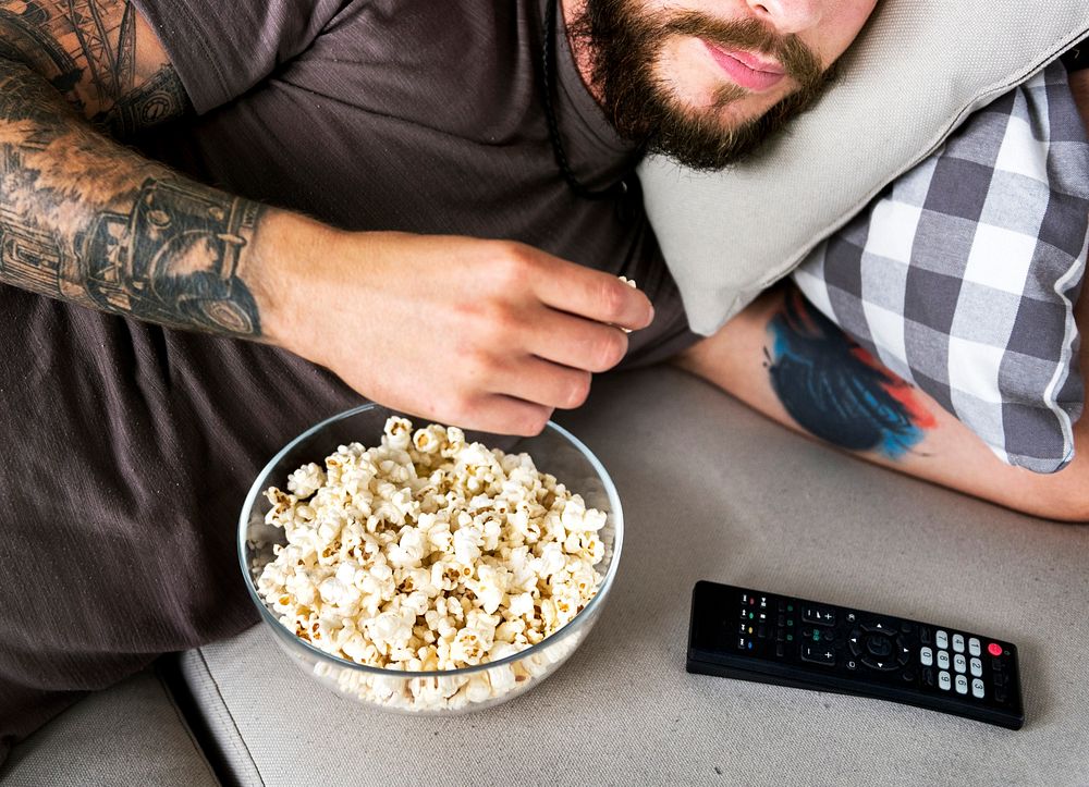 Man lying on a couch watching tv and eating popcorn