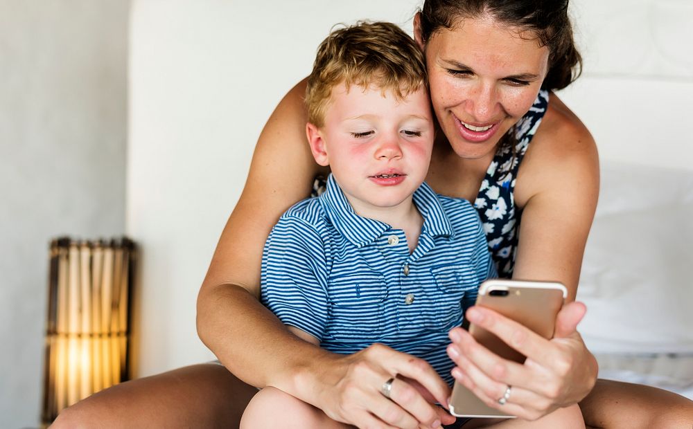 Mother sitting with son on bed using mobile