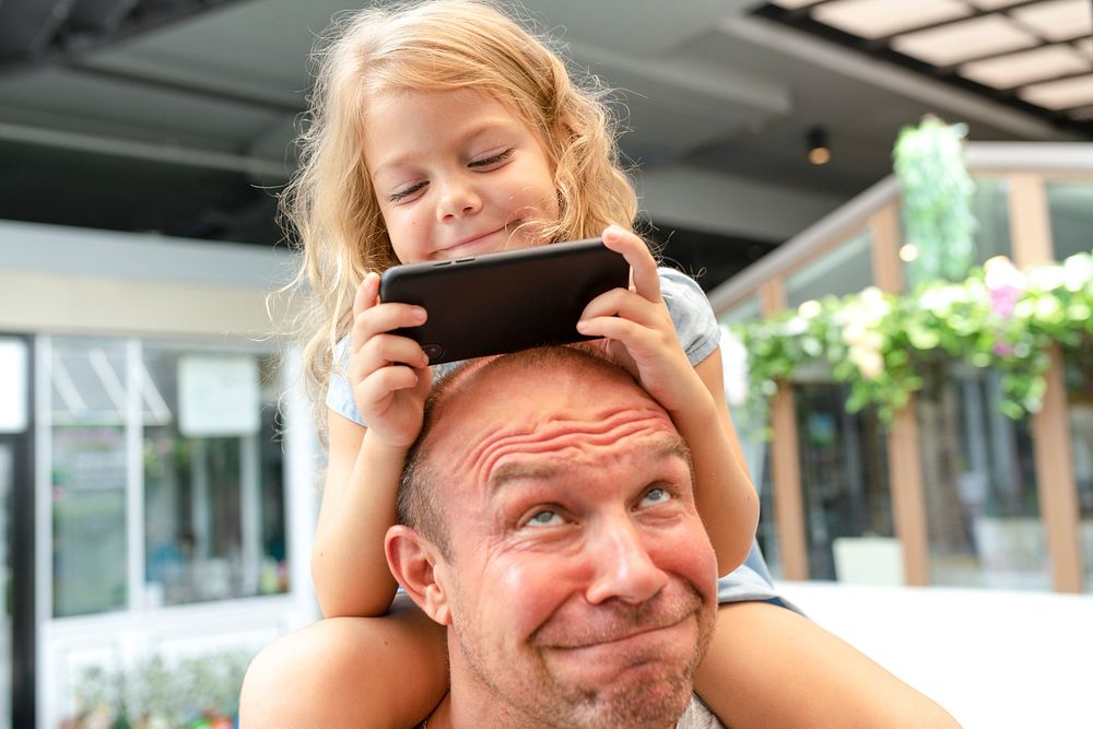 Little girl riding on her dad's shoulder and playing on a phone