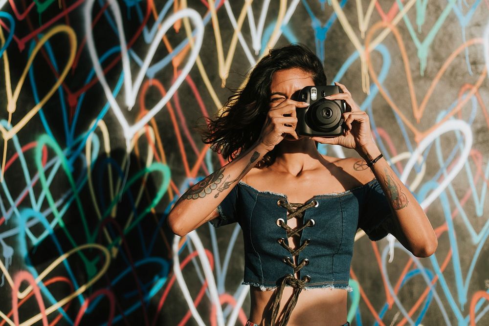 Cheerful woman using an instant camera against the backdrop featuring the graffiti artwork by James Goldcrown in Los…