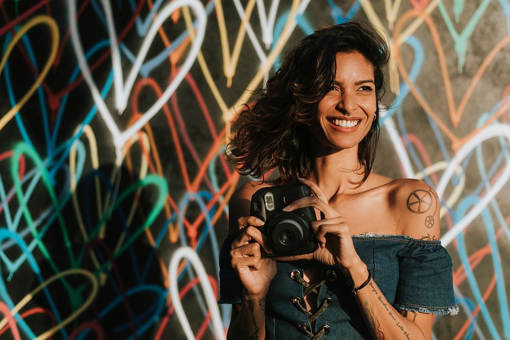 Cheerful woman using an instant camera against the backdrop featuring the graffiti artwork by James Goldcrown in Los…