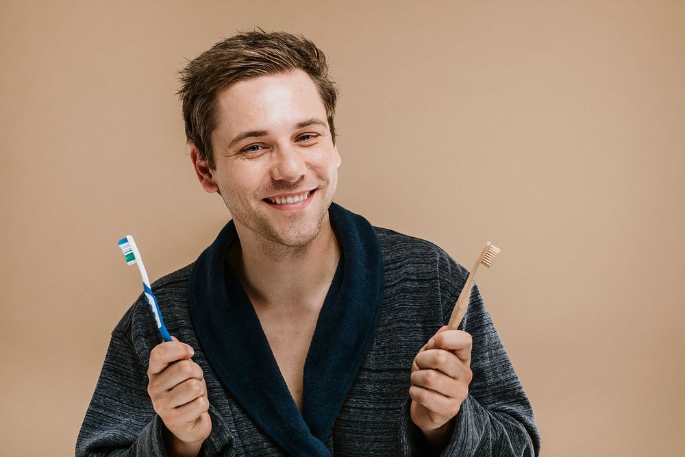 Blond man in a robe choosing between a wooden toothbrush and a plastic one