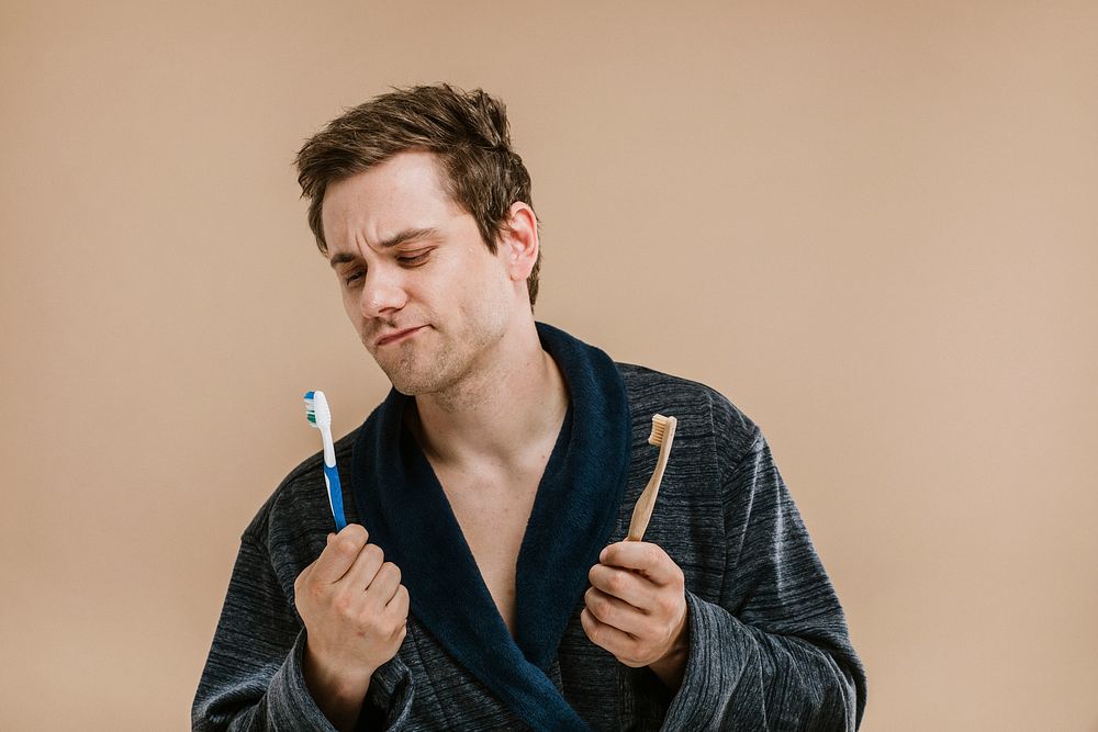 Blond man in a robe choosing between a wooden toothbrush and a plastic one