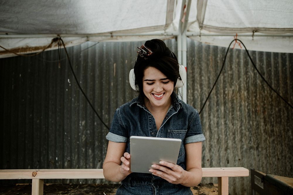 Cheerful female carpenter with protective headphones using a tablet at a workshop