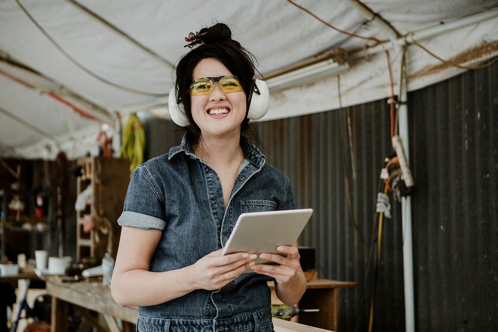 Cheerful female carpenter with protective headphones using a tablet at a workshop