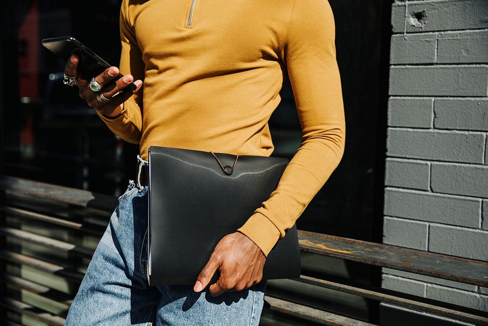 Man in a mustard yellow long sleeve top with a black folder briefcase using a phone