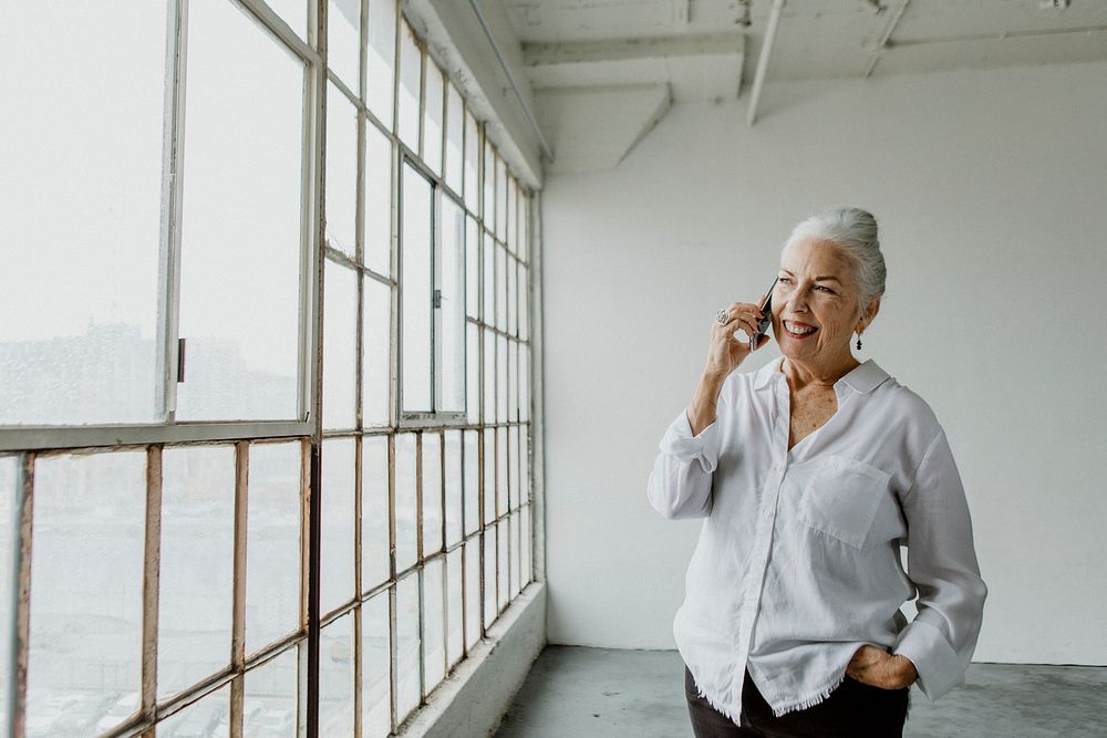Cheerful senior woman talking on a phone by the window in a white room