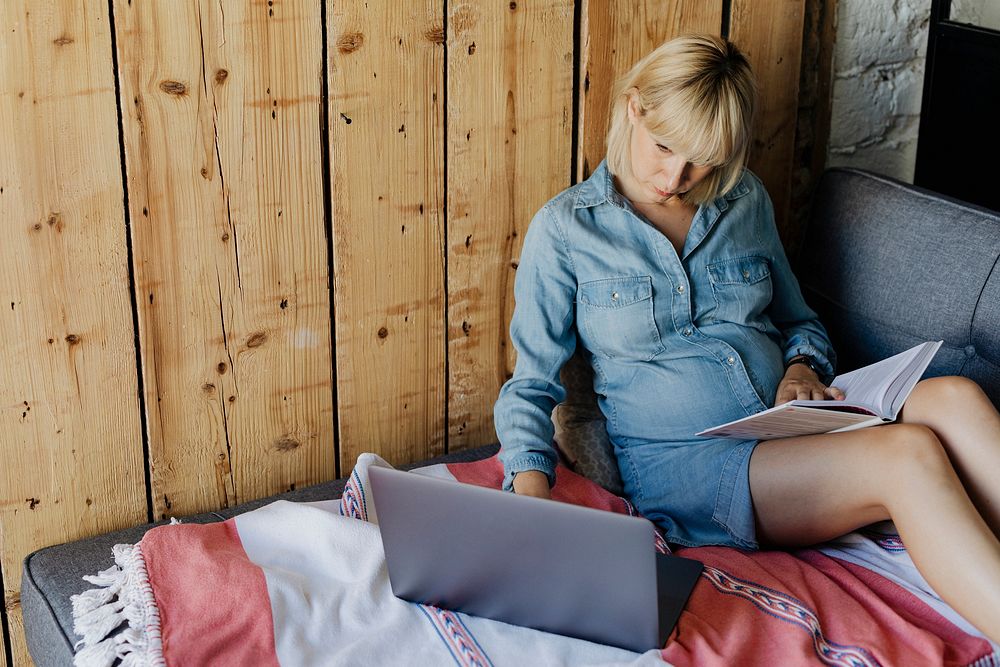Expecting mother reading a book while using a laptop